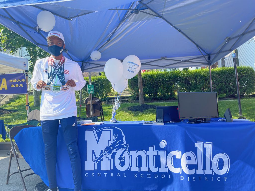 evan waterton is standing in front of a table with a blue tablecloth that reads Monticello. He is wearing a white shirt and has three medals draped across his neck. 