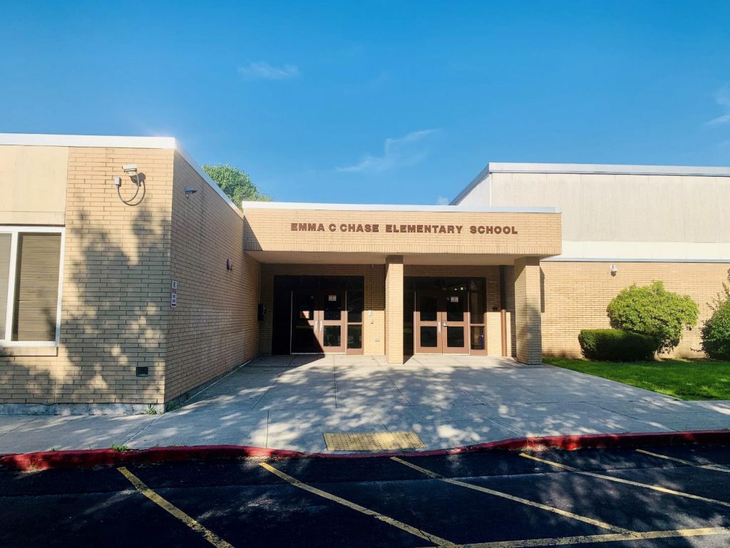 A tan brick building with the words Emma C Chase Elementary School written on top. There is a parking lot in front and a blue sky above.