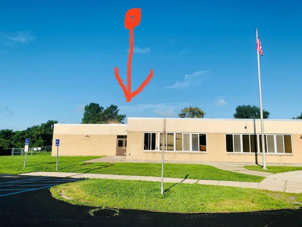 A light tan brick building, with green grass in front, blue sky above, and a flagpole on the right. There is a red arrow pointing to a door on the left.