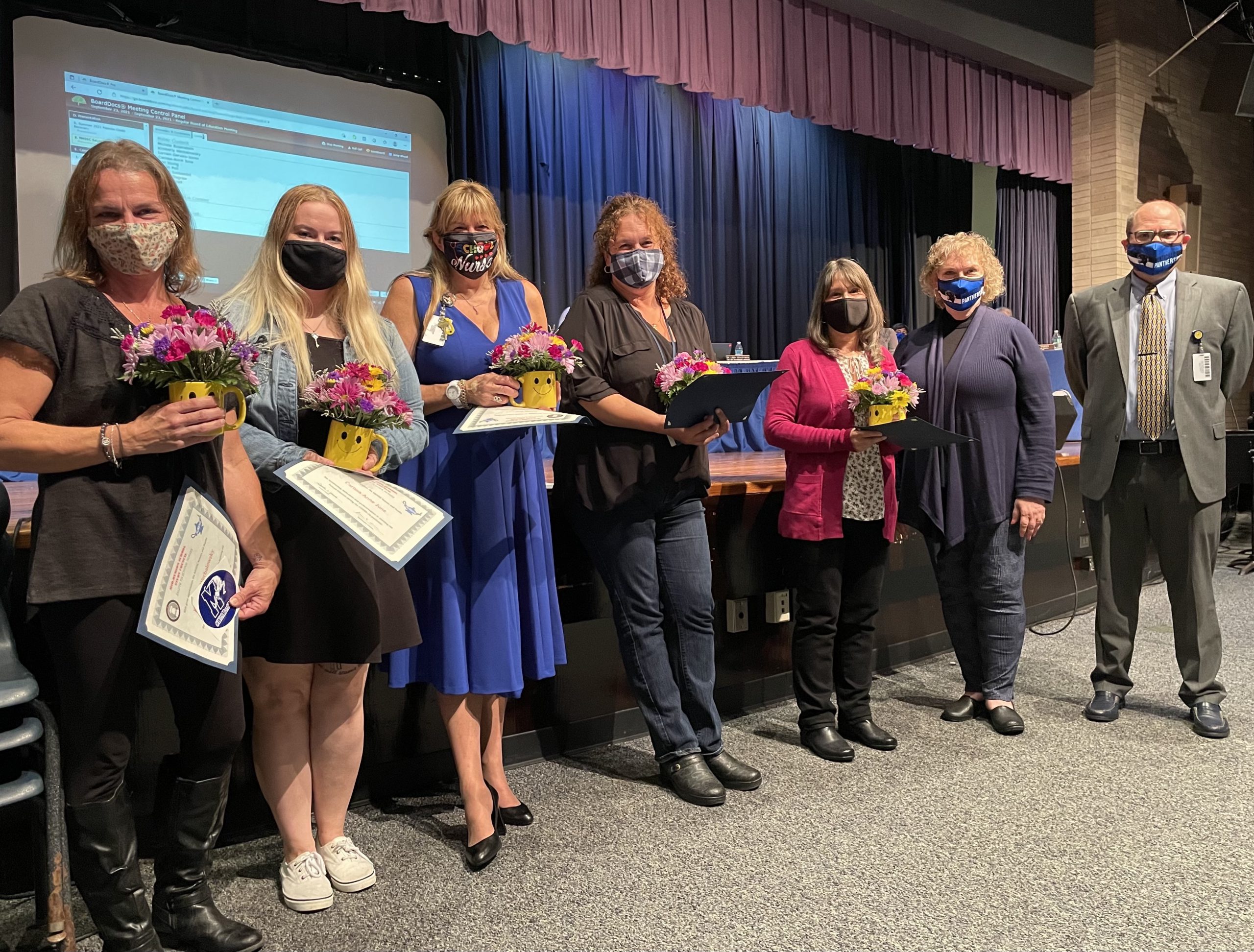 a group of school nurses standing in front of the high school auditorium and holding flowers. They are flanked by Board President Lori Orestano James and Superintendent Matt Evans