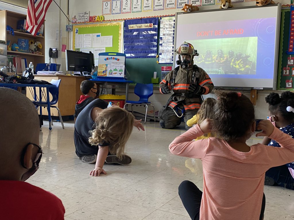 Deputy Michael Barrett is seated on the floor in fire fighter gear, speaking with children 