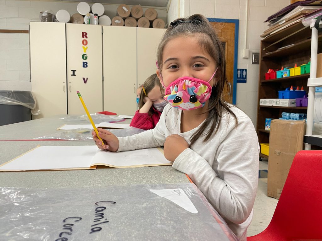 a young girl with brown hair in a ponytail is seated at a desk. She is wearing a mask and smiling