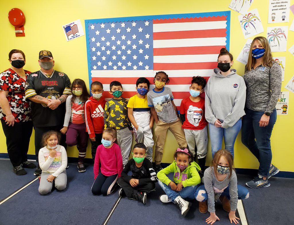 Ms. Dutcher and Miss D’Agata’s first grade class with Retired US Marine Corps Sergeant Joseph Kavanaugh standing in front of the USA Flag bulletin board that the students helped to measure and create. 