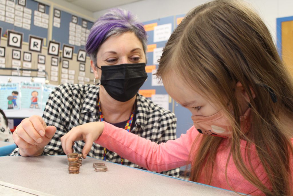 a teacher and student are seated at a desk. The student is counting coins as the teacher watches