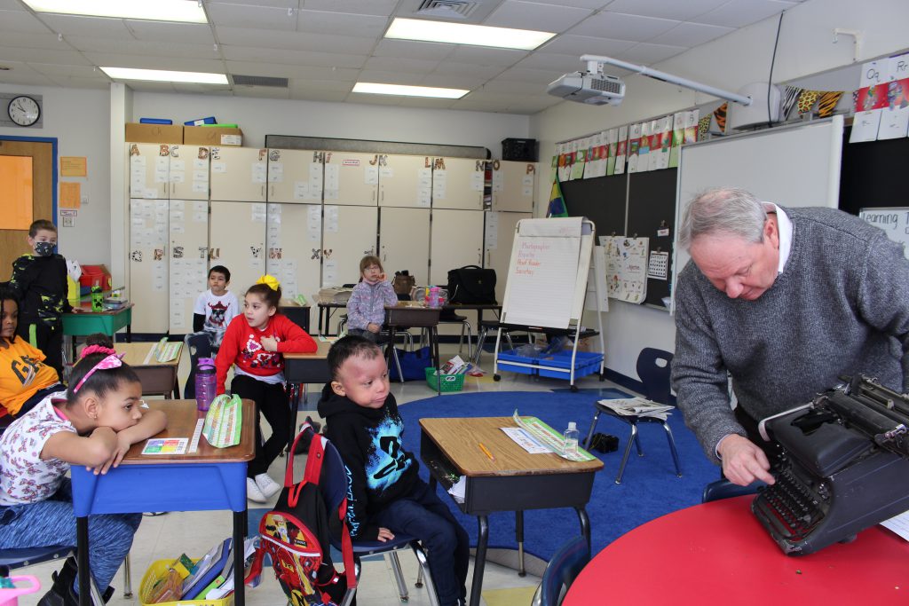 Fred Stabbert is demonstrating how to type on a type writer. A classroom of children is watching