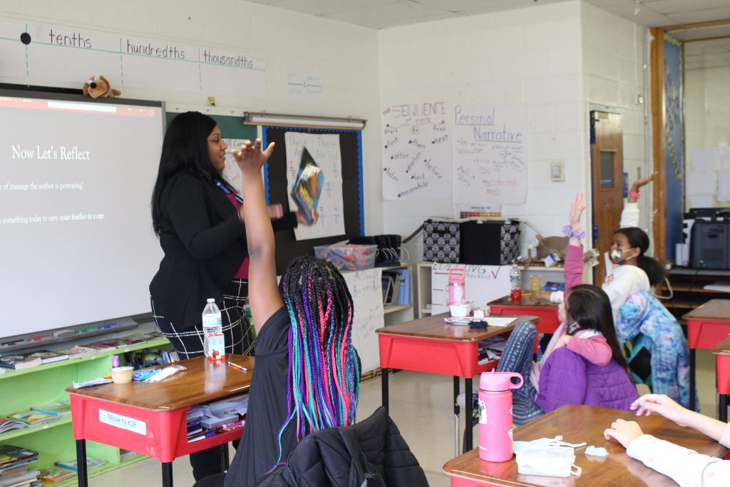 the director of student equity is standing at the front of a classroom, asking a question. Three students have their hands raised, poised to answer. 