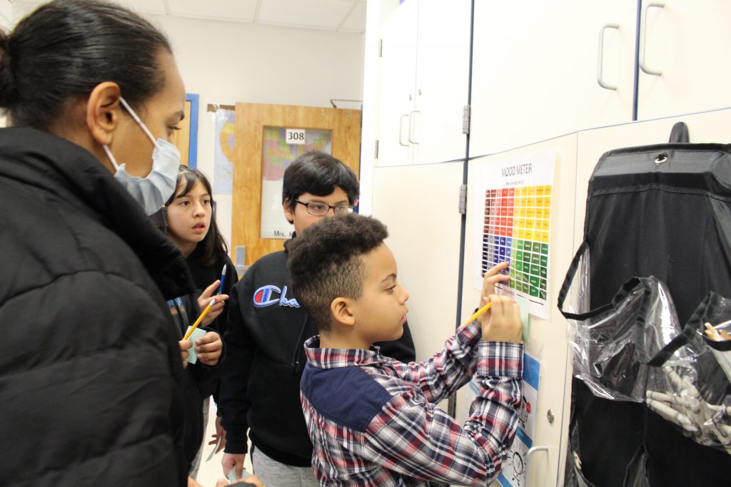 a group of students is huddled around a poster that says "mood meter." They are reading the mood meter to help them figure out what to write on their post it notes. 