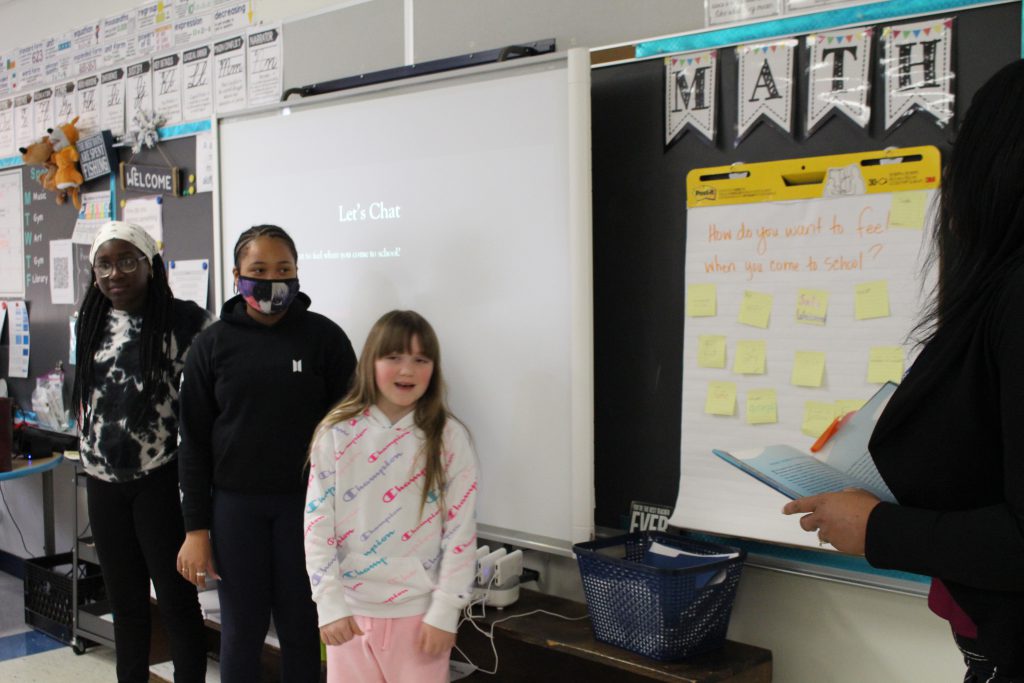 three students are standing at the front of a classroom speaking to the class