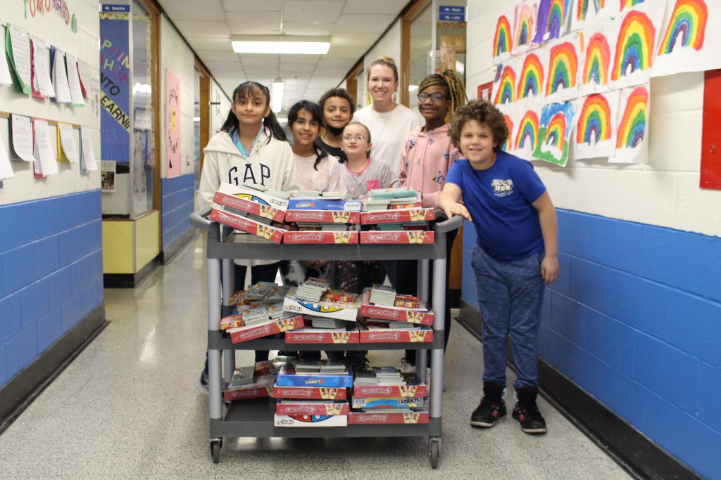 a group of students is standing with their teacher next to a cart. On the cart, there are goody bags 