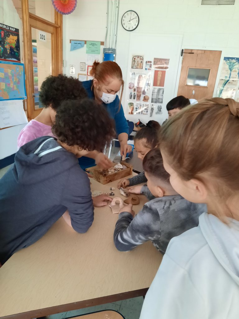 Ms. Reef is showing students some of the tiles. The students are crowded around a table looking down at the mosaics. 