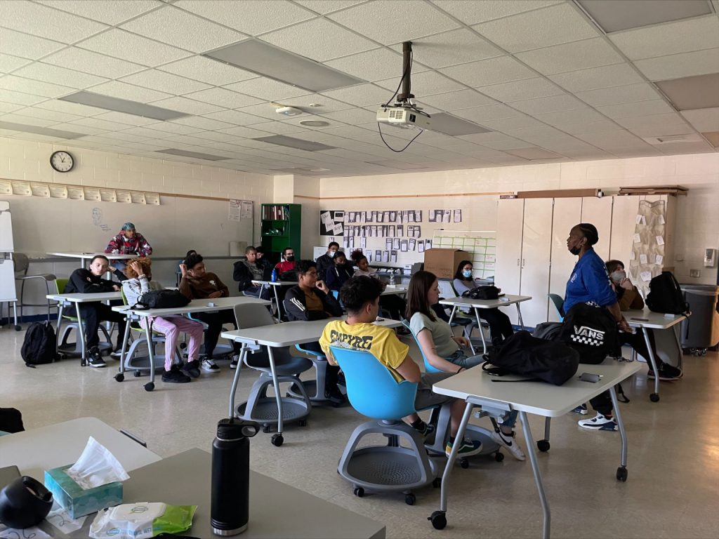 A woman in a blue t-shirt is addressing a classroom filled with high school-aged students