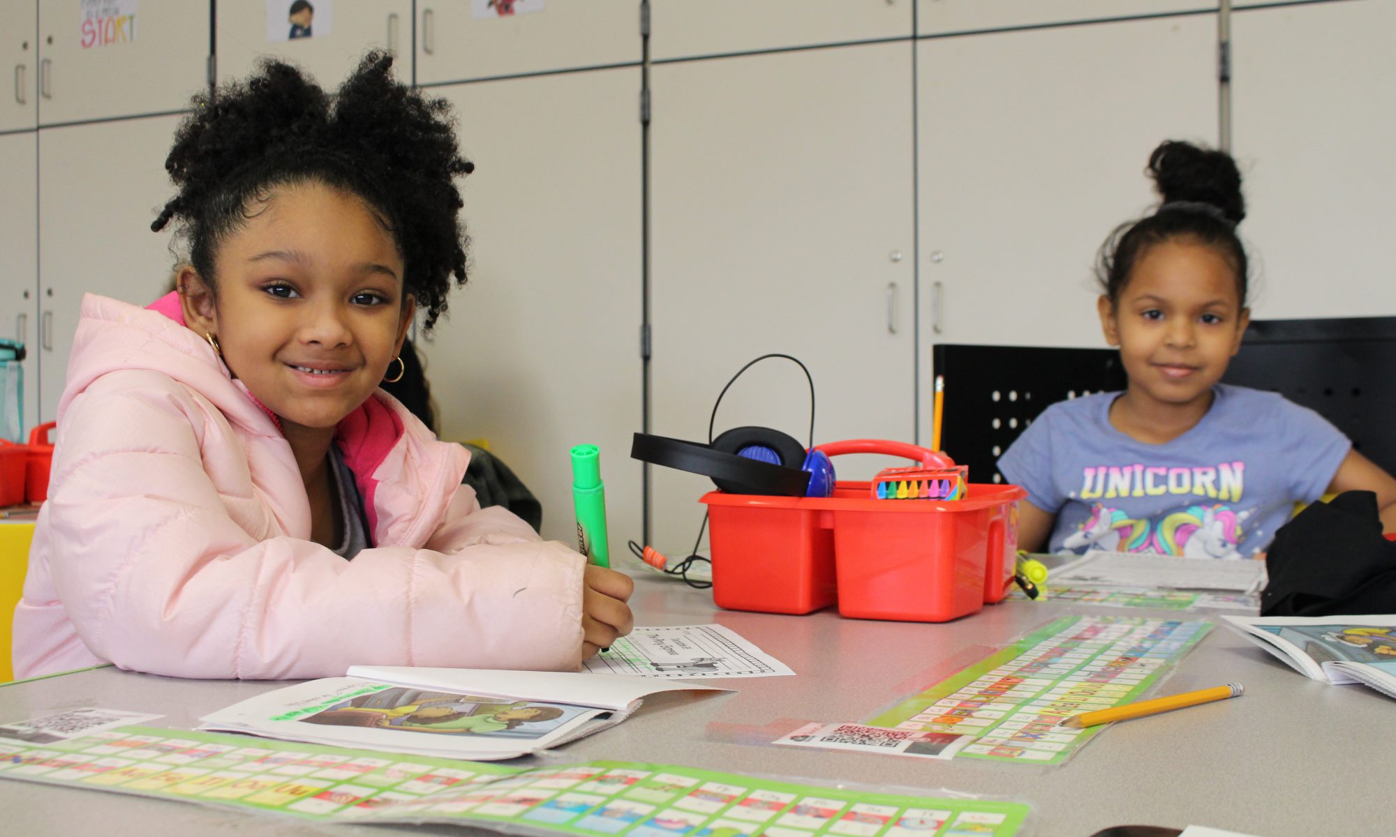 two girls are seated at a desk and smiling at the camera