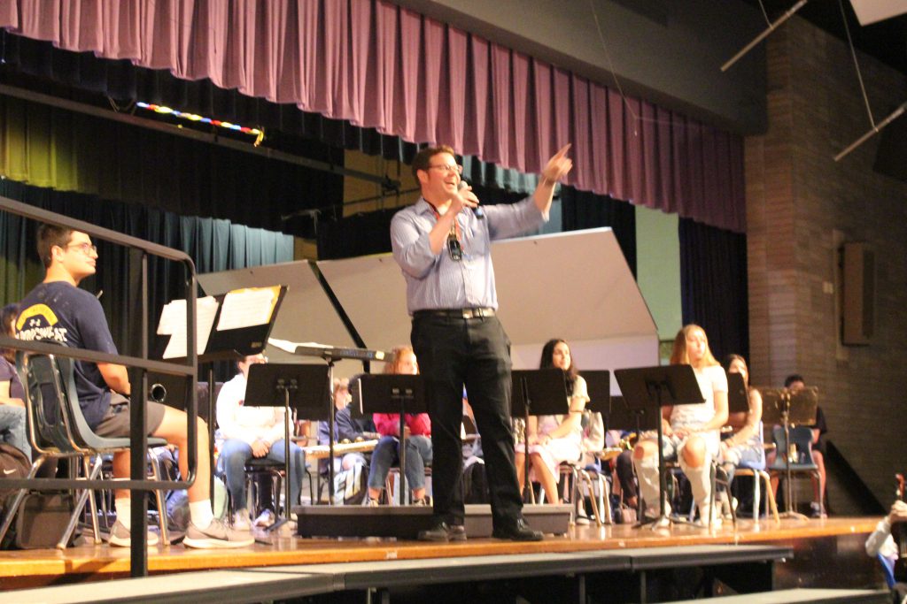 A student band is onstage and a man with a microphone is standing in the center adressing the audience 