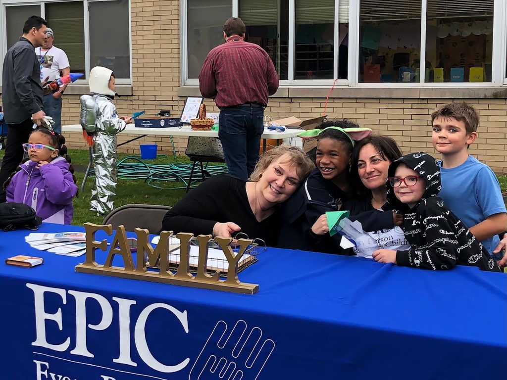 Jane Sorensen and Lori Orestano-James are sitting at the blue EPIC table outside Cooke Elementary smiling for a picture with 3 elementary students.