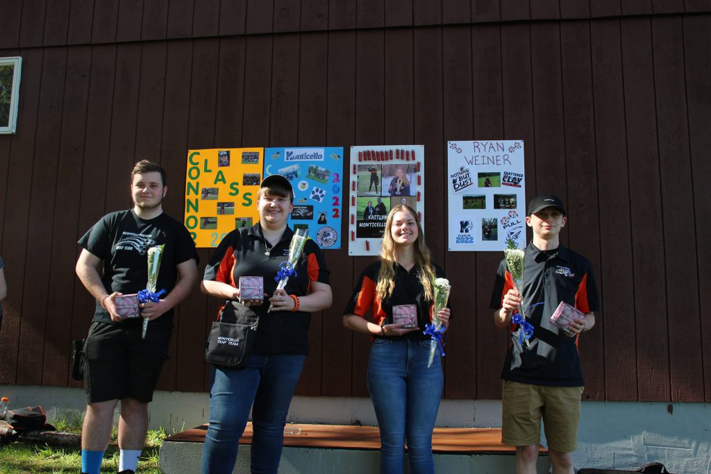 four students are posing and smiling holding medals. Behind them are posters saying congratulations