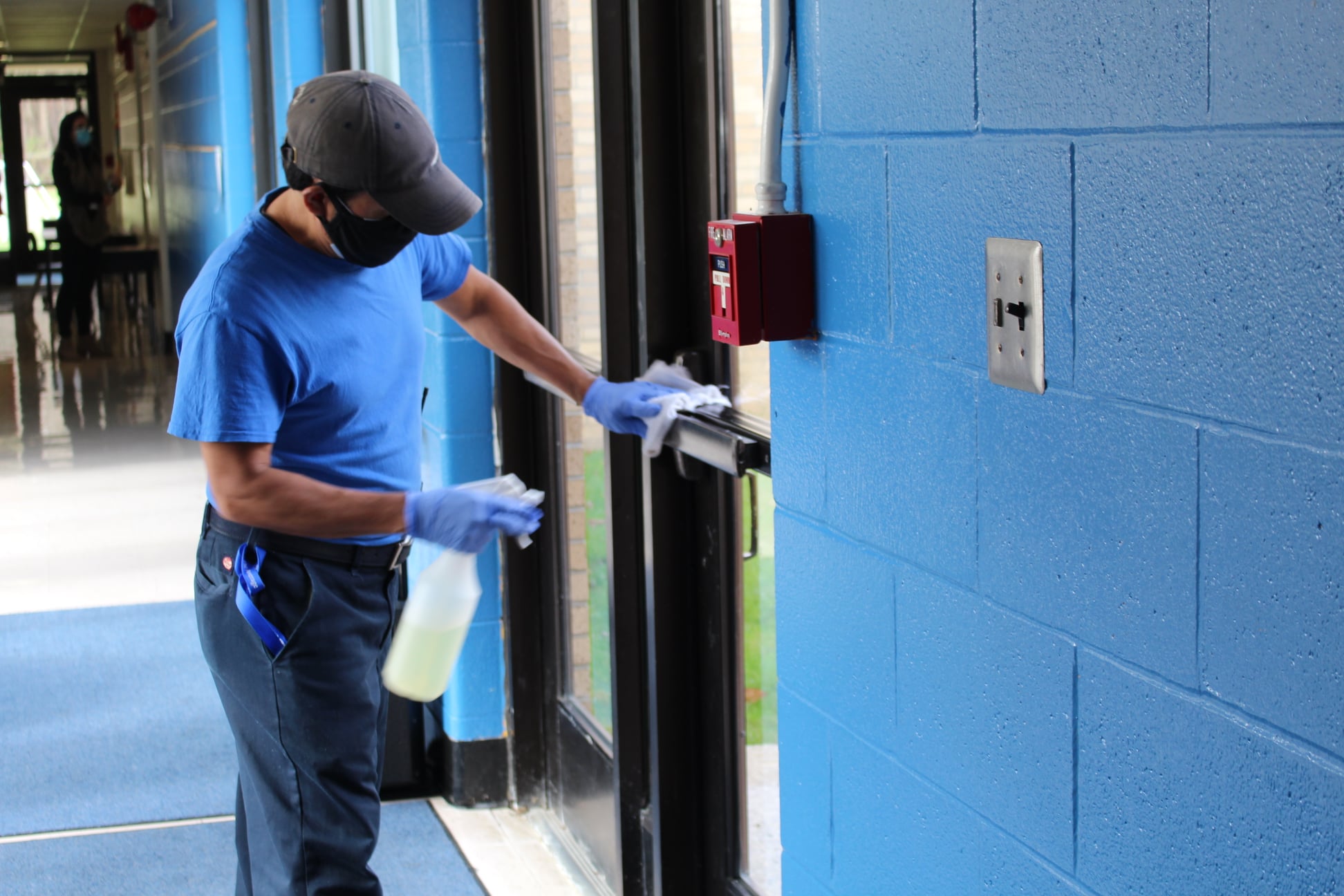 A custodian is cleaning a door handle. He's wearing a face mask and holding a cloth and spray bottle.
