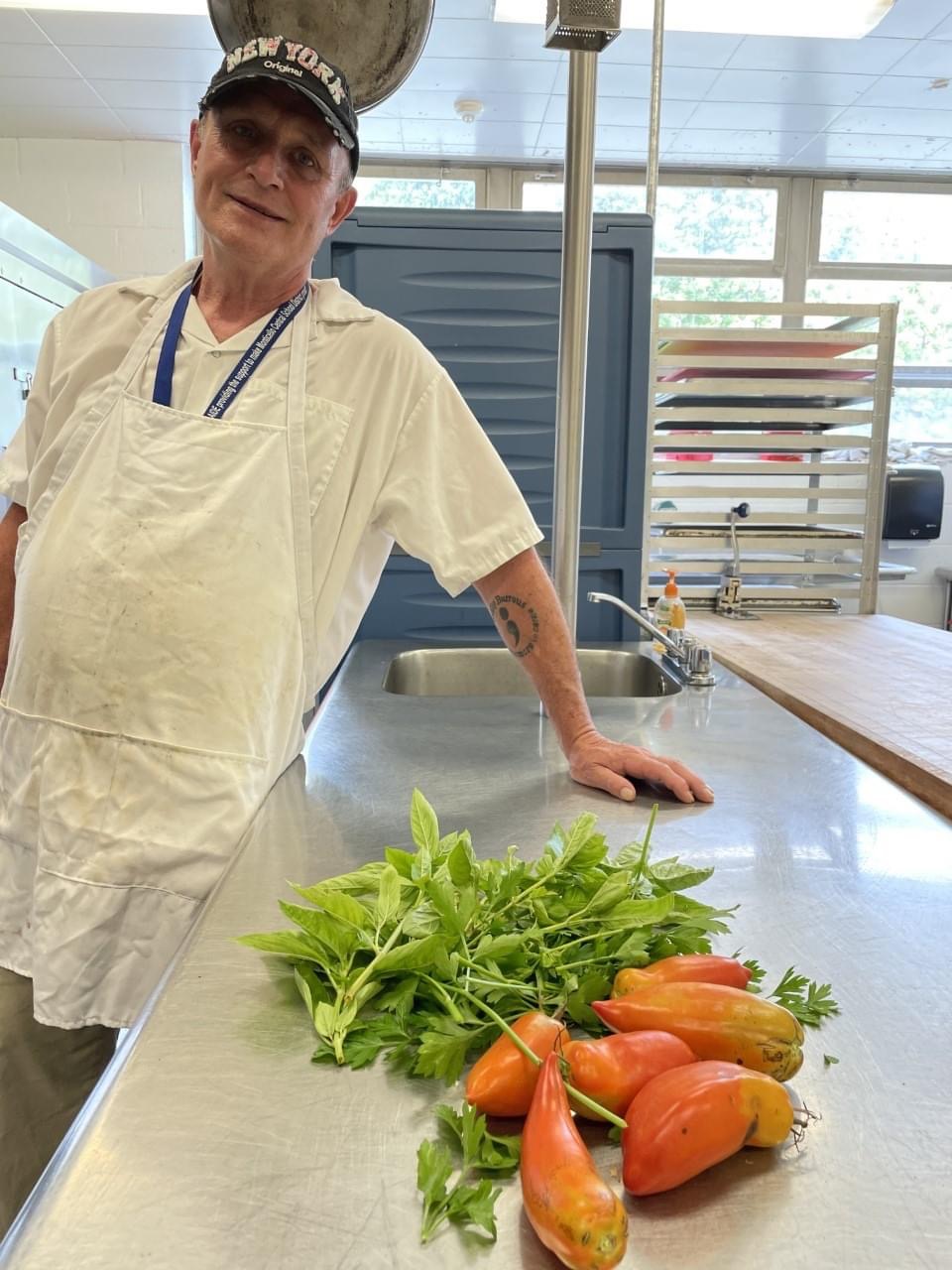 A male food service worker is smiling for a picture in front of a counter with vegetables on top of it.