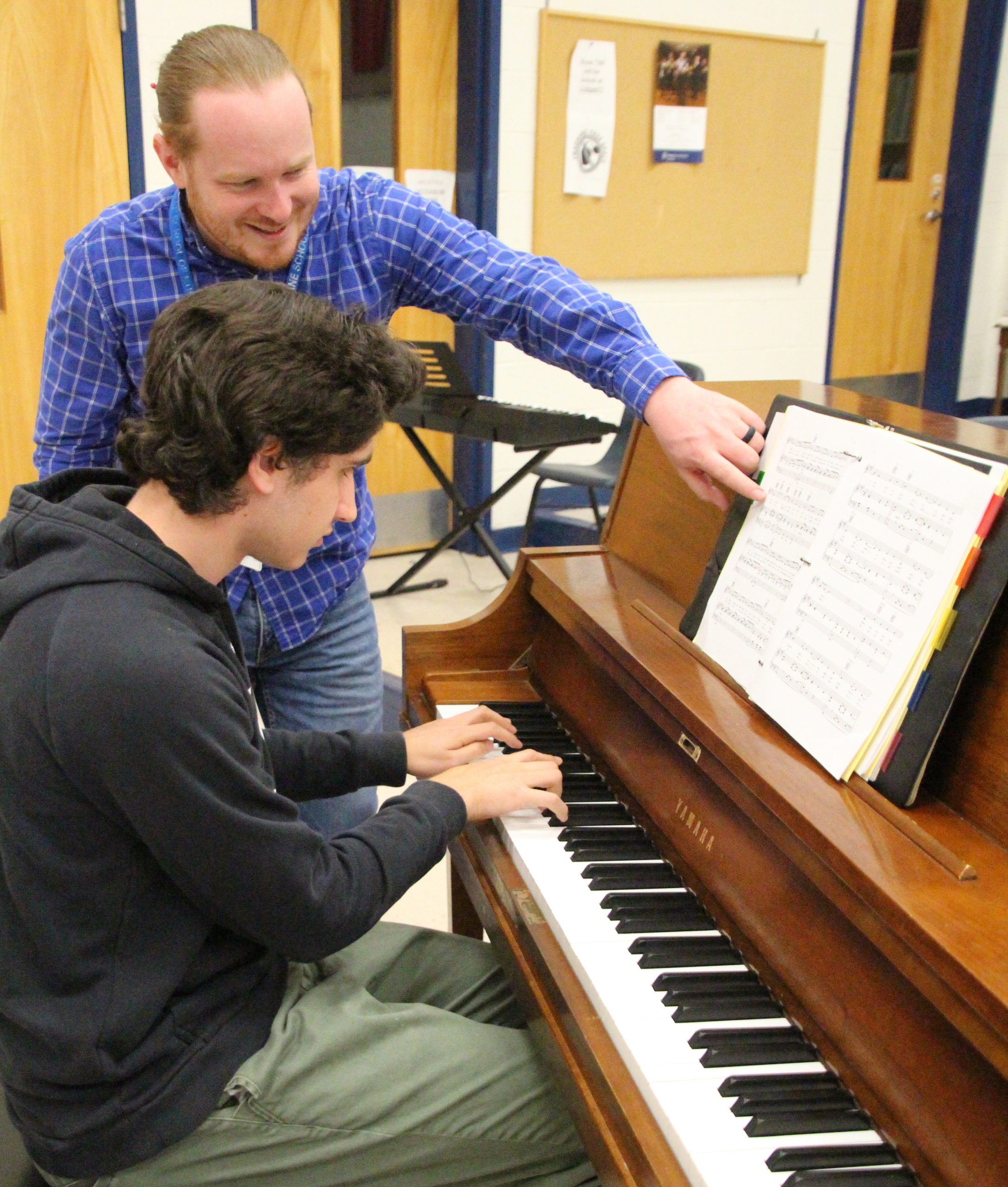 A music teacher is helping a student who's playing the piano. The teacher is pointing to the sheet music in front of the student.
