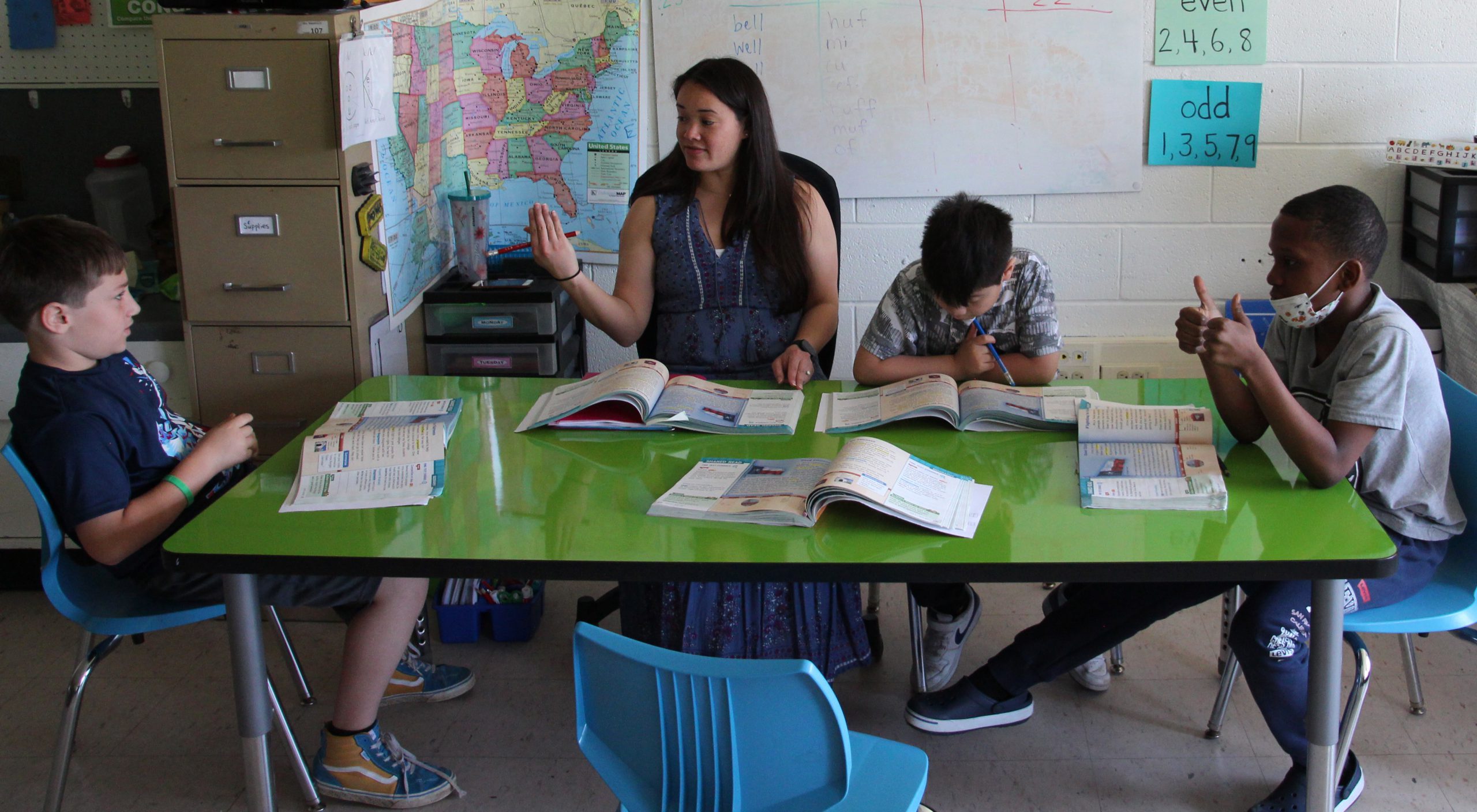 A teacher is sitting at a table with 3 students. Textbooks are open in front of everyone and the teacher is engaging in conversation with the student on her left.