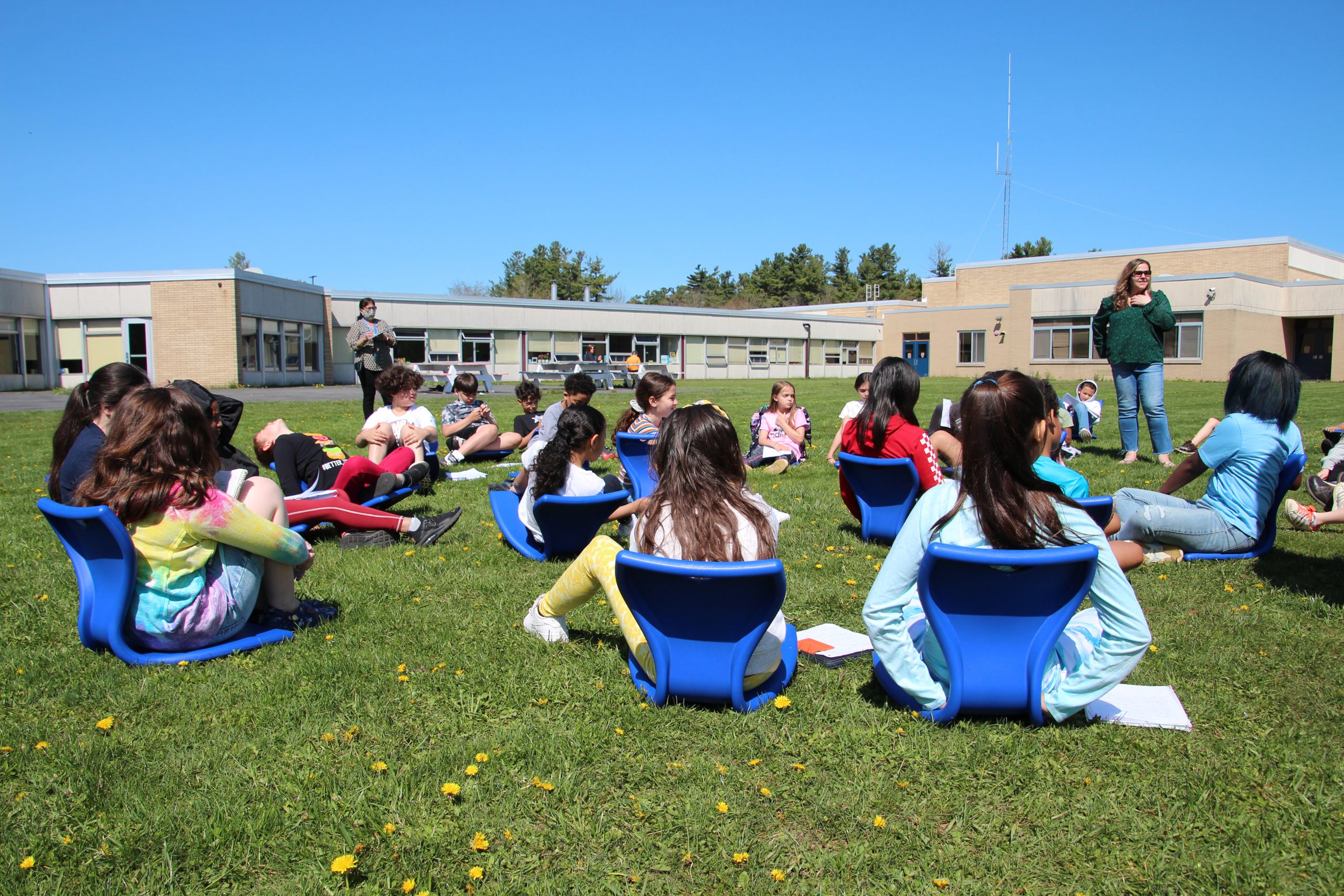 A class is sitting outside in blue chairs on the grass. They're watching their teacher as she speaks.
