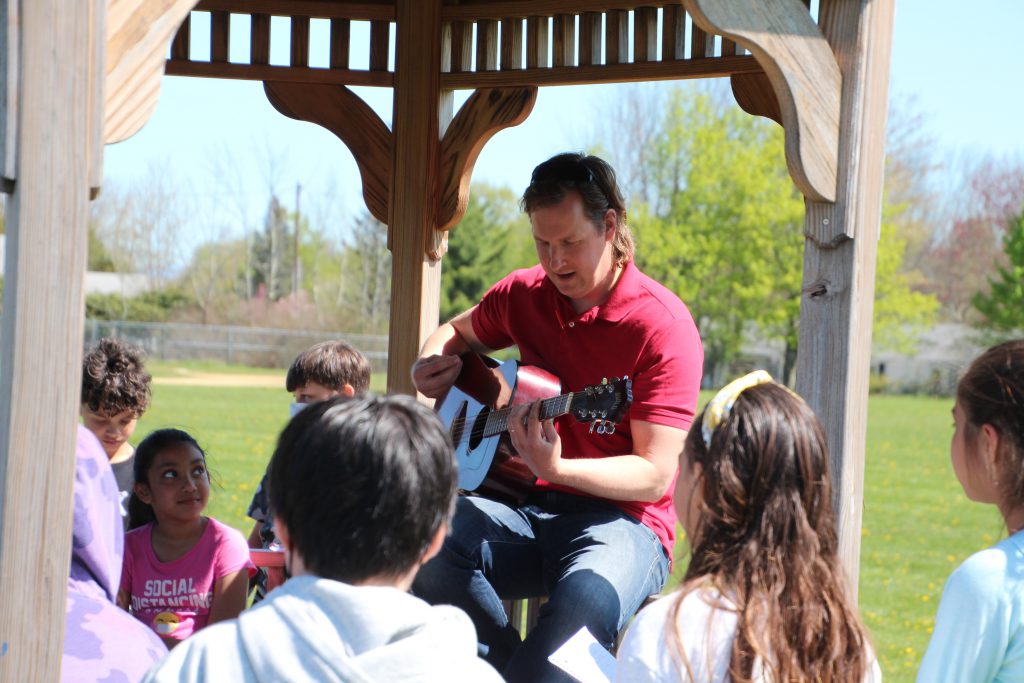 A music teacher is playing the guitar for his students in the gazebo outside of the school.