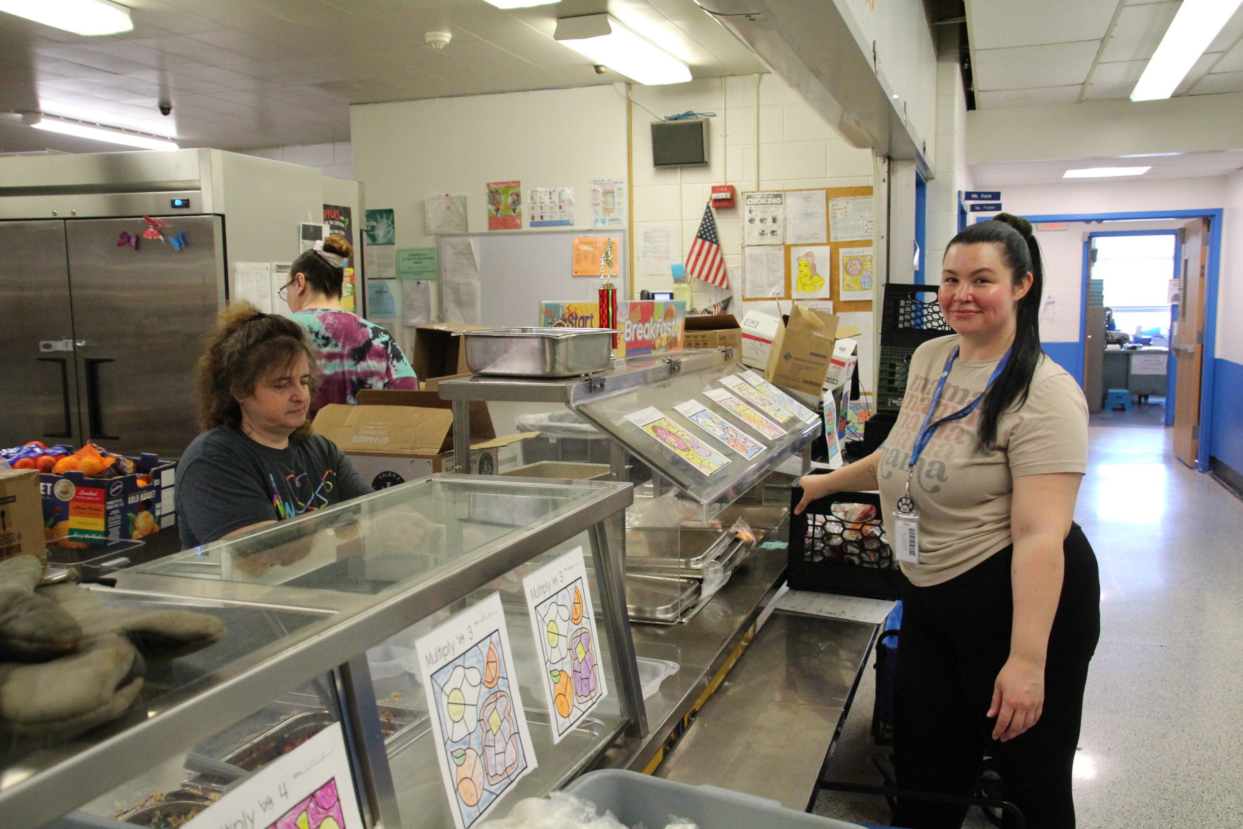 A food service worker is looking at the camera and smiling for a photo in front of the kitchen. Another food service worker is working behind the counter.