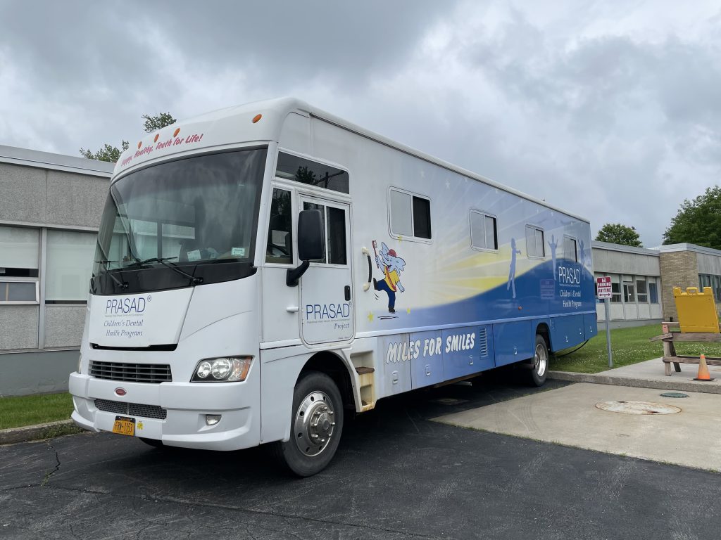 the PRASAD dental van is parked outside of a school building. It is the size of a Winnebago and says PRASAD on the side