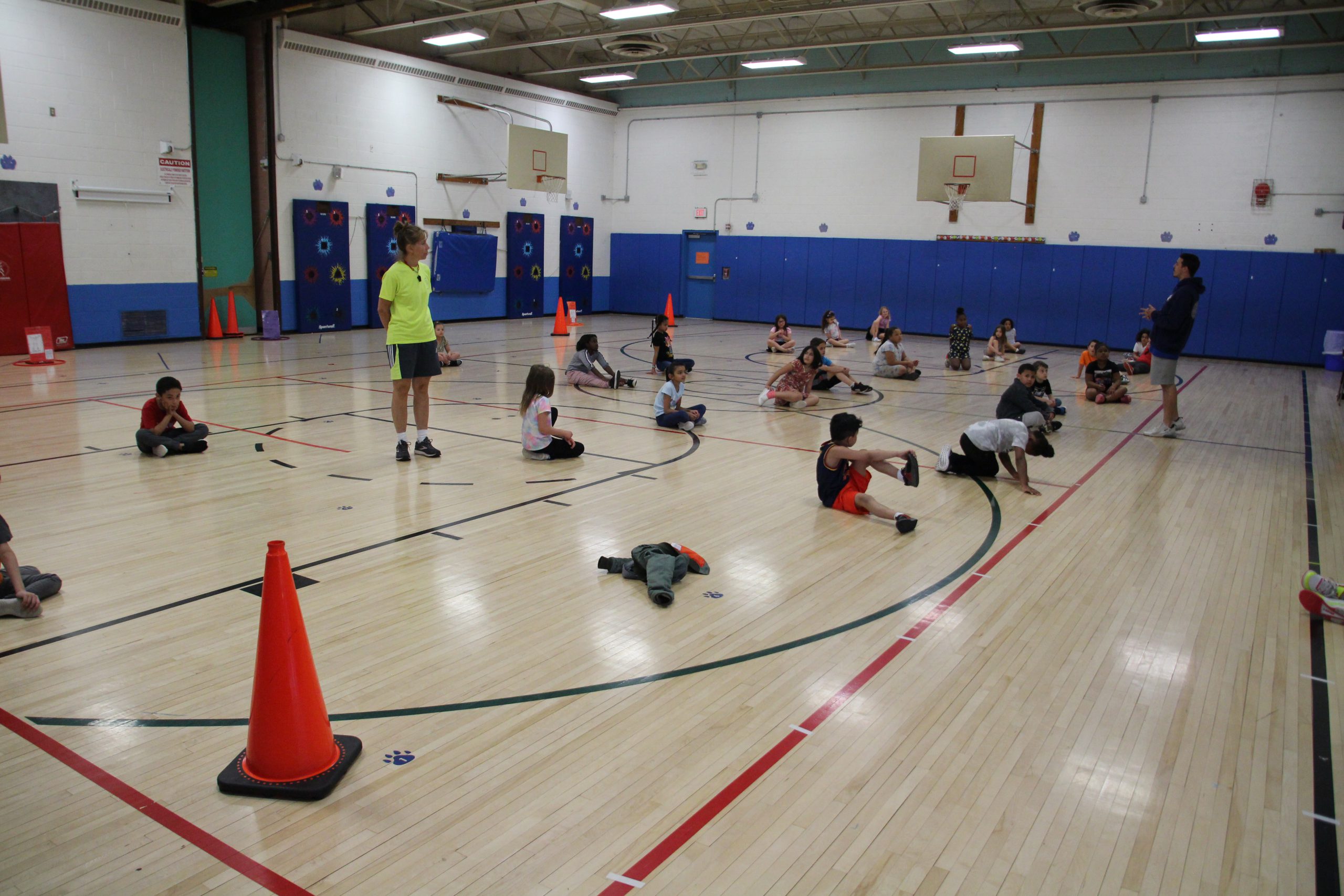 A Monticello staff member wearing a yellow tee shirt is standing on the court while students are on the ground doing an exercise.