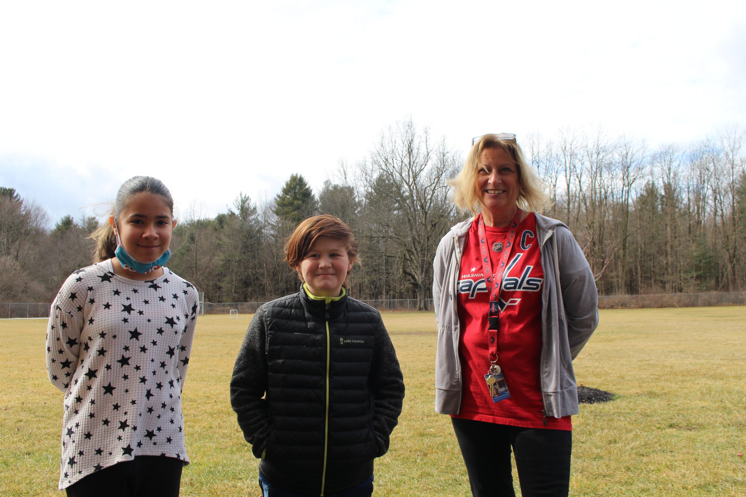 Maryann Swensen is standing with two of her elementary students outside. The field can be seen in the background.