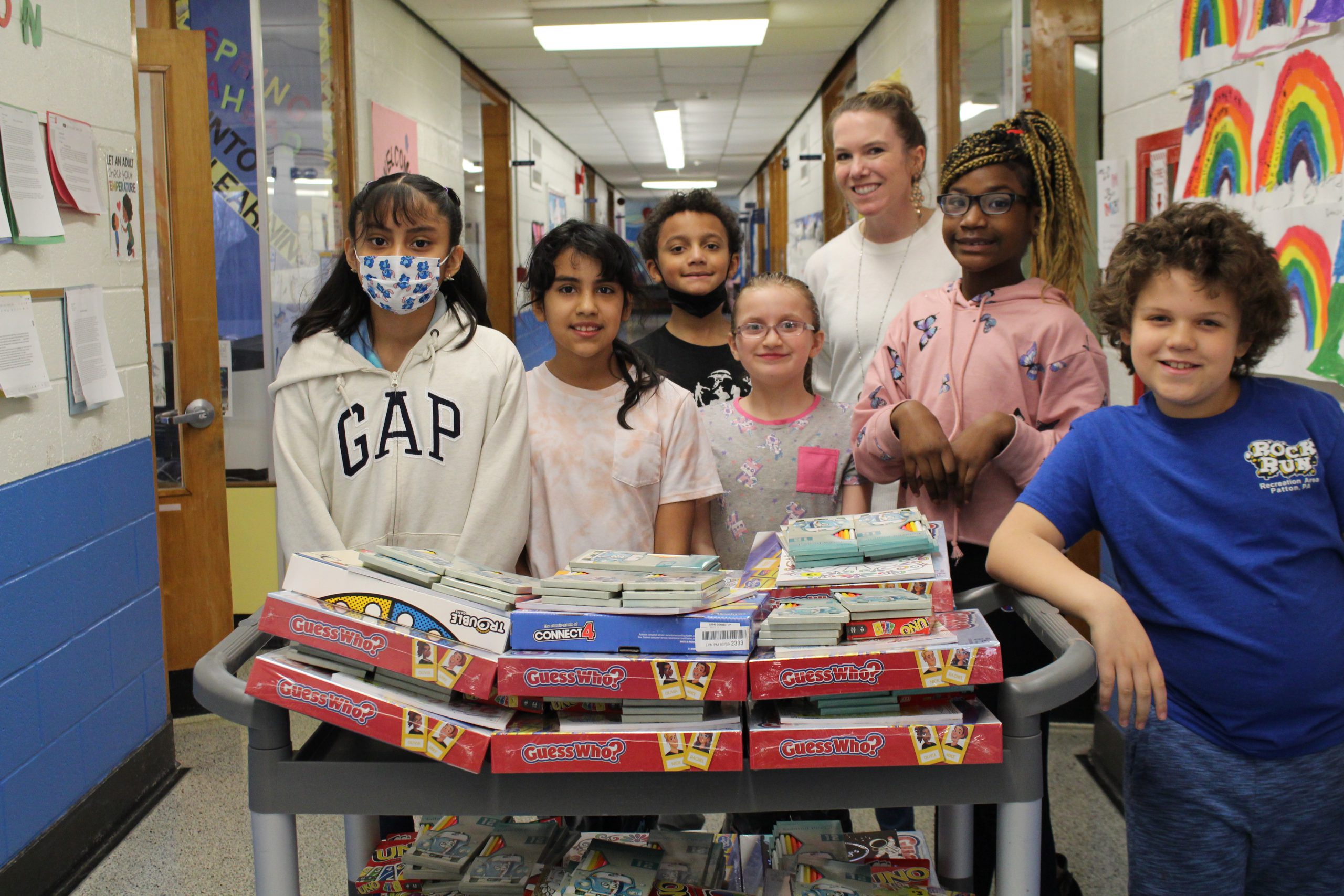 Monticello students and a Monticello staff member are standing in a hallway in front of a cart filled with board games. Everyone is standing together and smiling for a picture.