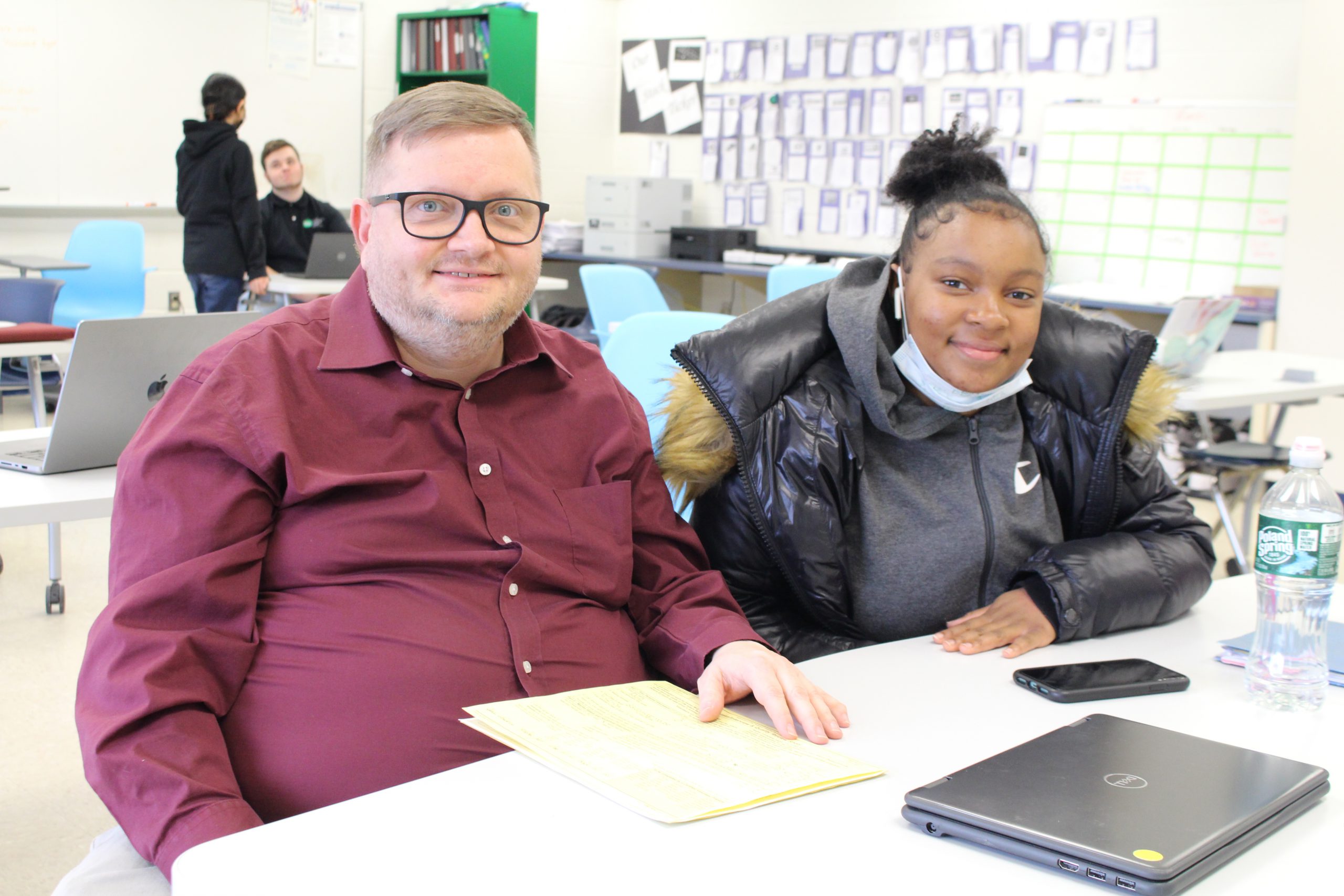 A teacher is sitting next to a student at a table and both are smiling for a picture.