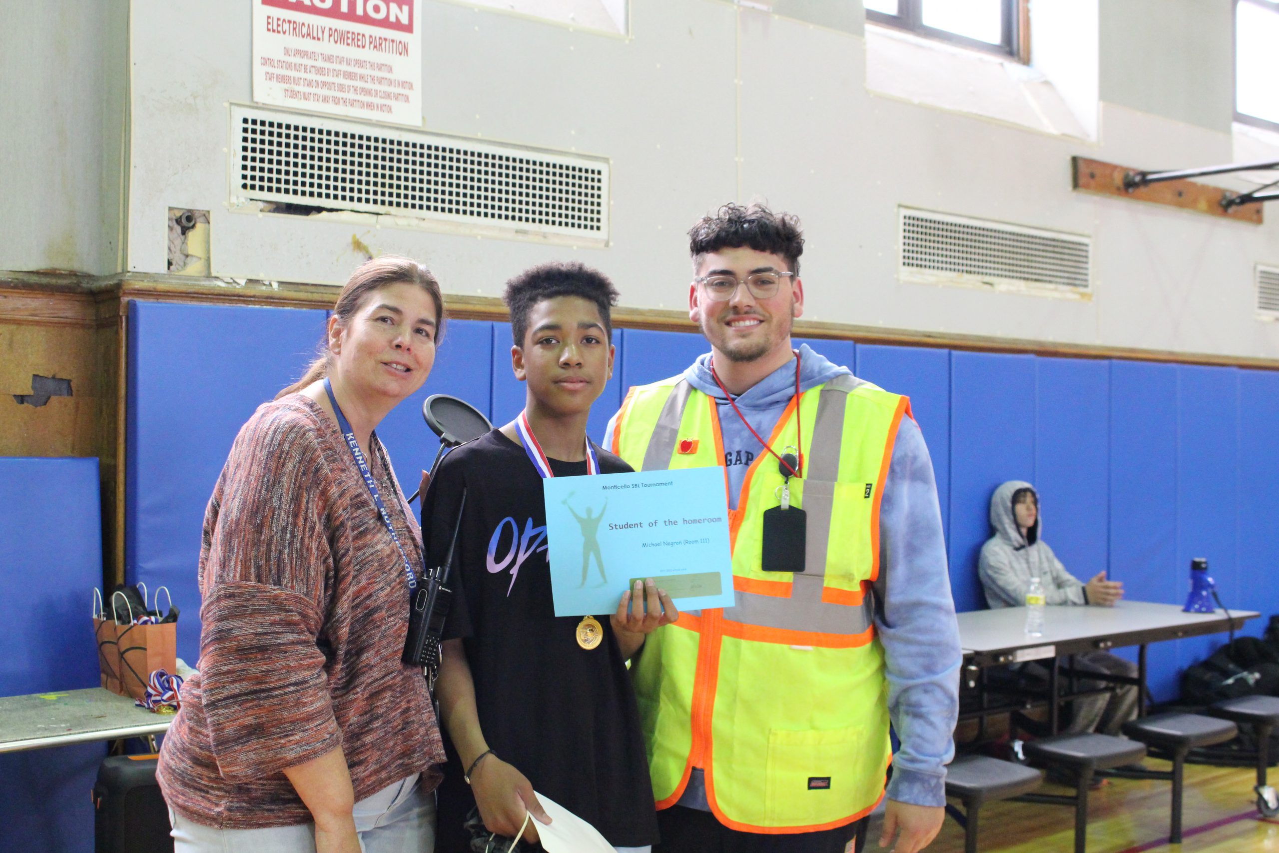 A Monticello staff member with a walkie-talkie around her neck is standing with a security attendant (who's wearing a bright yellow reflective vest) and a student. Everyone is smiling for a picture as the student holds up a certificate.