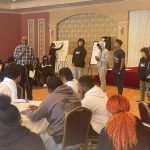 a group of students are at the front of a ballroom addressing another group of students who are seated