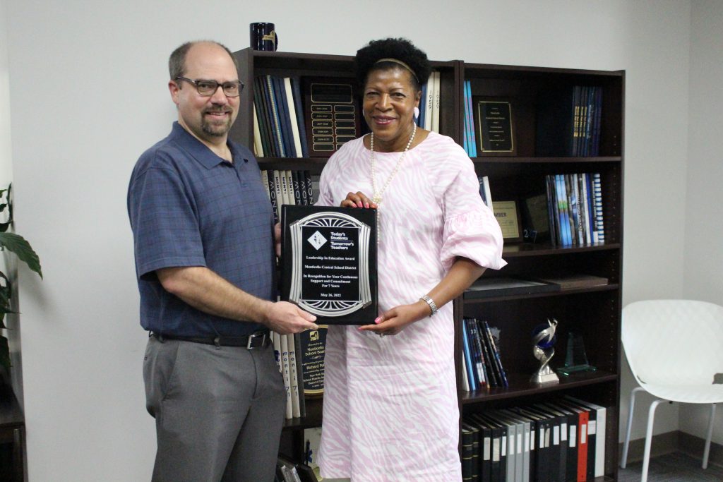 the superintendent of schools and the CEO of Todays Students Tomorrows Teachers are standing by a bookshelf and holding an award