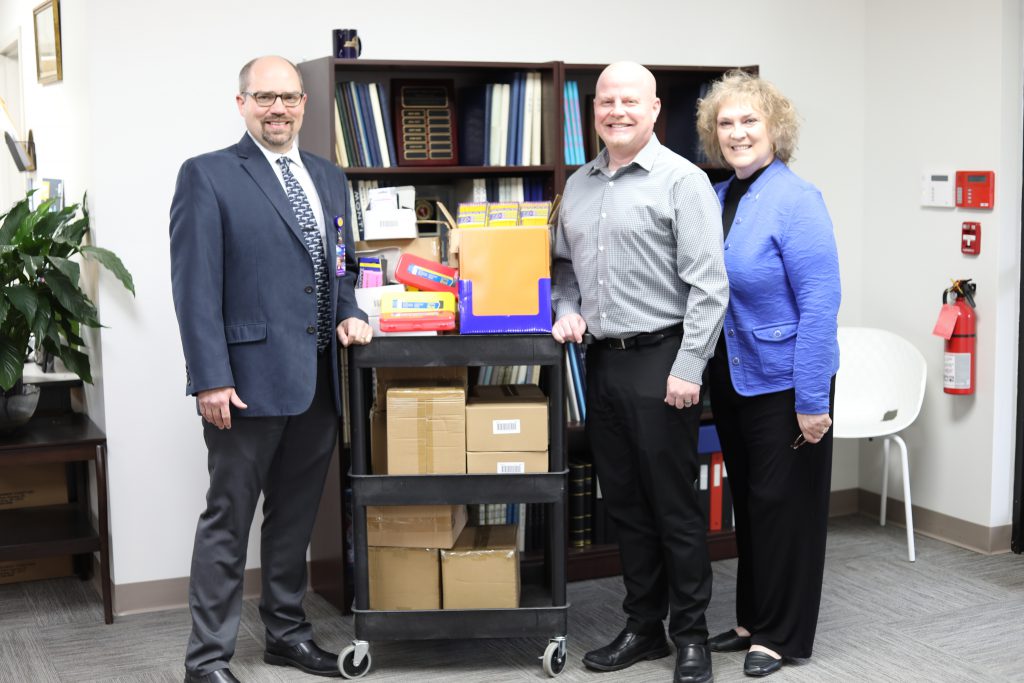 three people are standing around a cart filled with school supplies. 