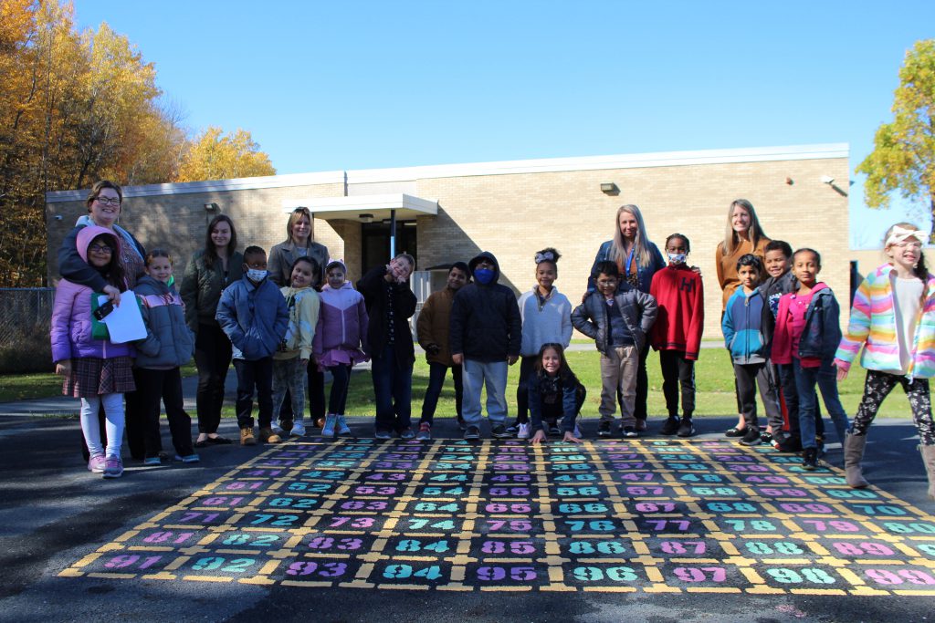 students in ms. dutcher and d'agata's class are posing by the number chart. The number chart is a painted chart on the asphalt ground of the playground. Students are standing around it. 
