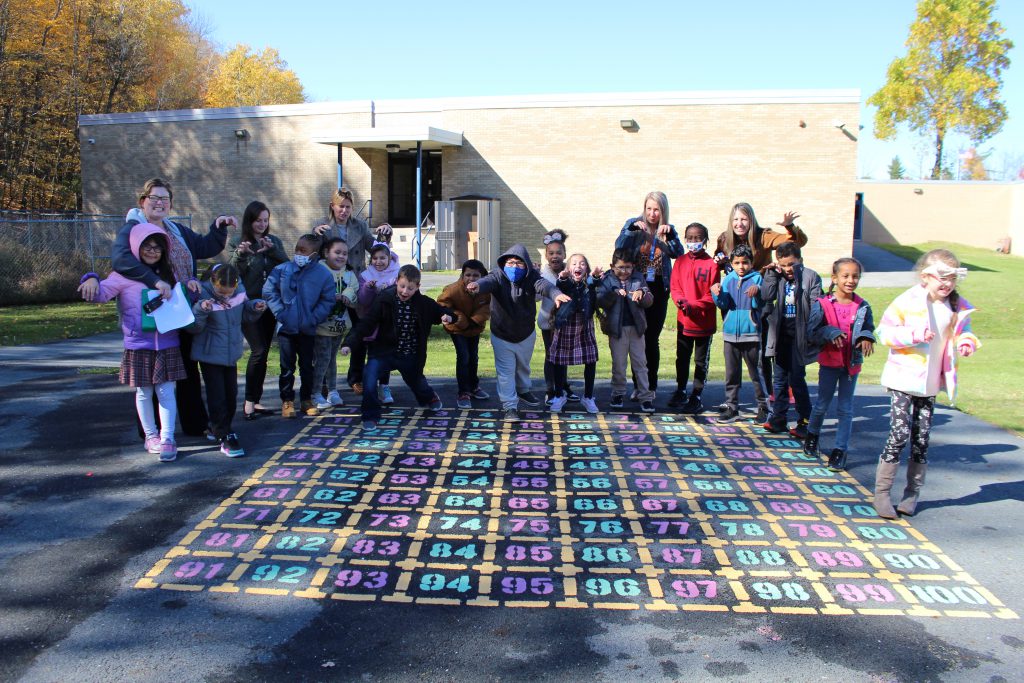 the classes are standing around the numbers chart. They are all posing with their arms out imitating a dinosaur.