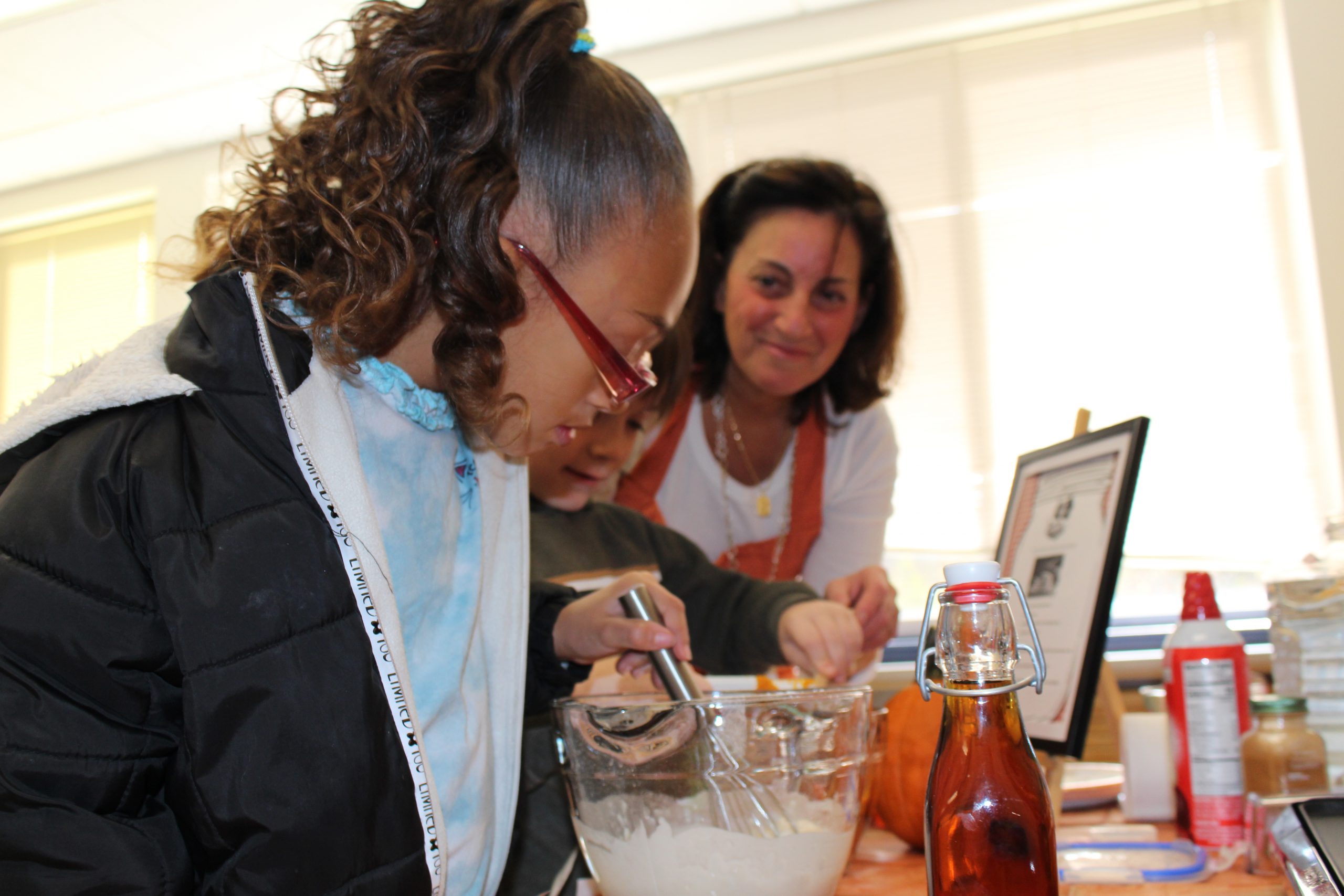 two young girls are mixing ingredients together at a table as a smiling woman looks on