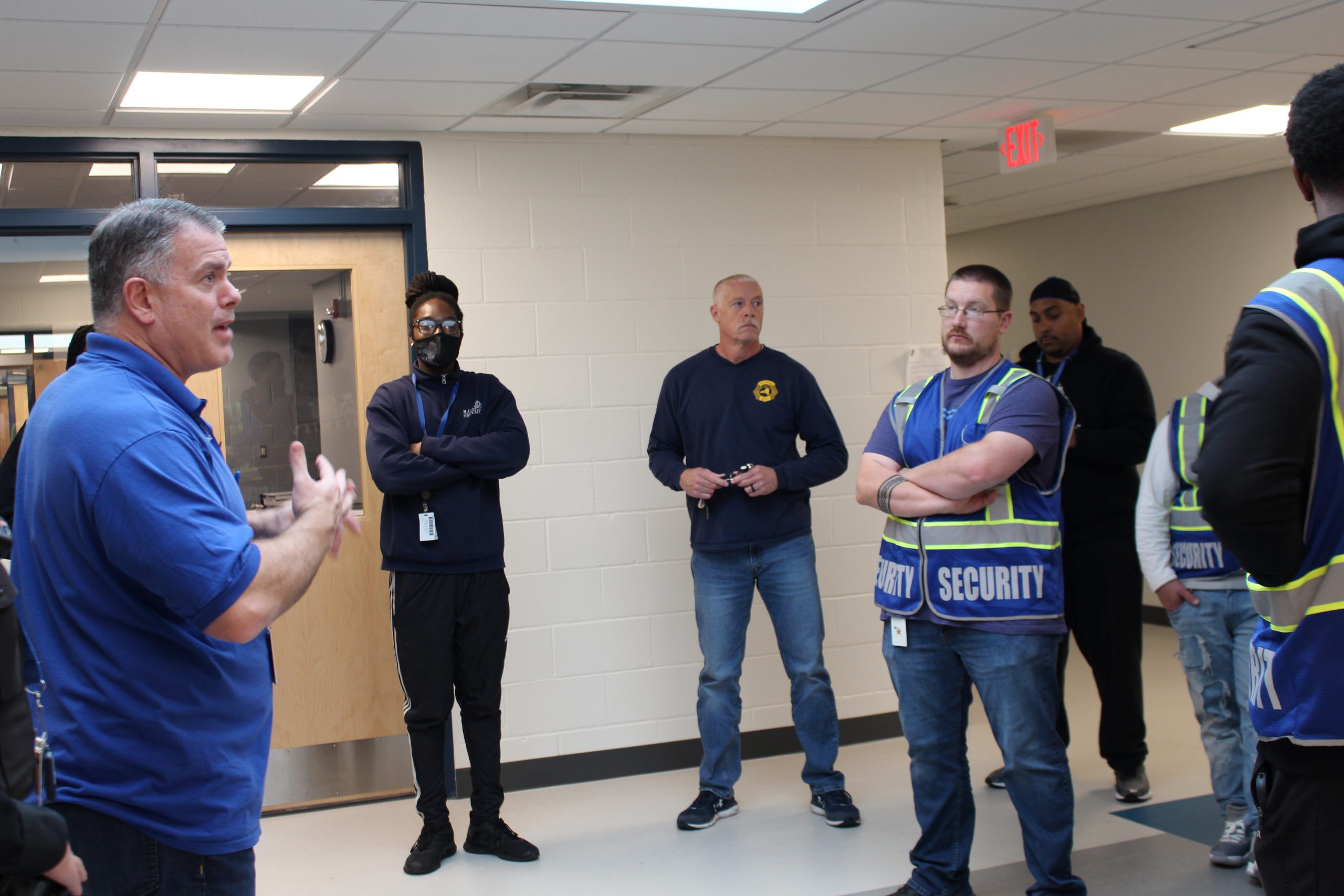 a group of security guards are standing in a circle. A man is in the center speaking to them 