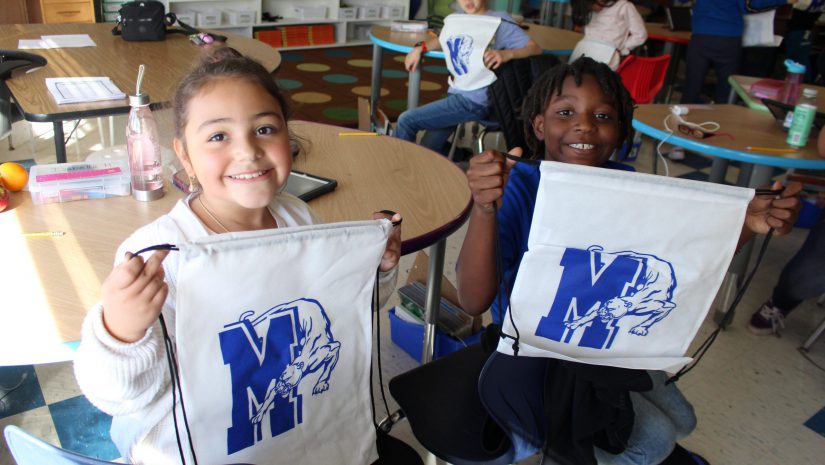 two students, one male and one female are holding up white bags with a blue Monticello panther logo and smiling.