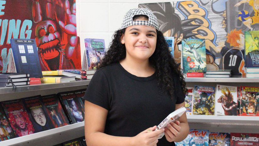 a pre-teen aged girl with long curly dark hair is holding a book and smiling at the camera