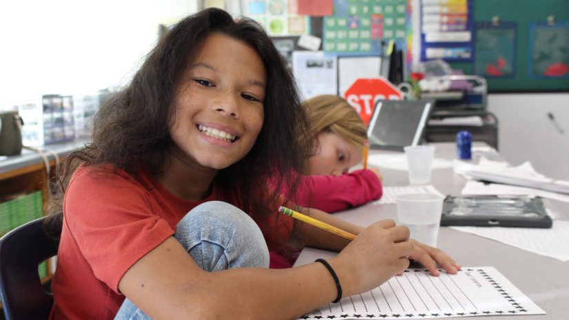 a student is seated at a desk and writing. He is wearing an orange shirt and is smiling up at the camera.