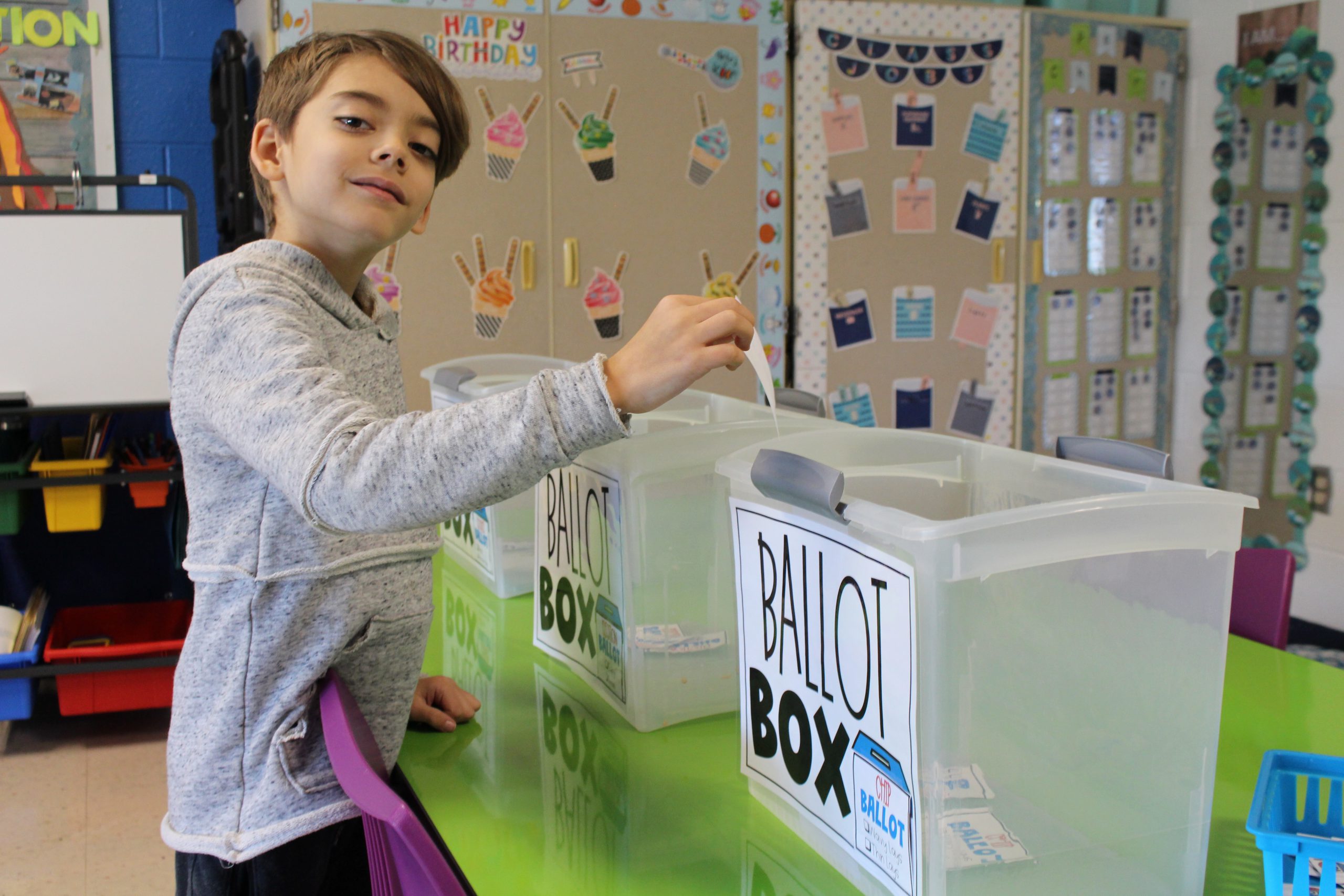 a boy is smiling. He is about to drop a ballot into a box that says "ballot box"