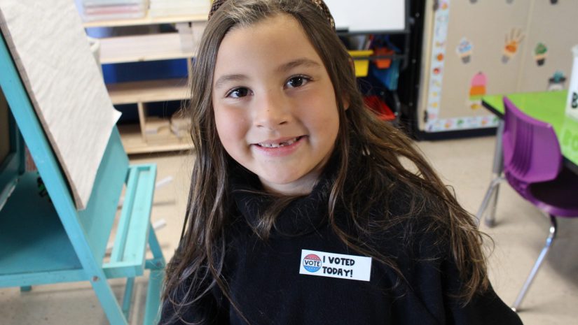 a young girl is smiling at the camera. She is wearing a sticker that says "i voted"