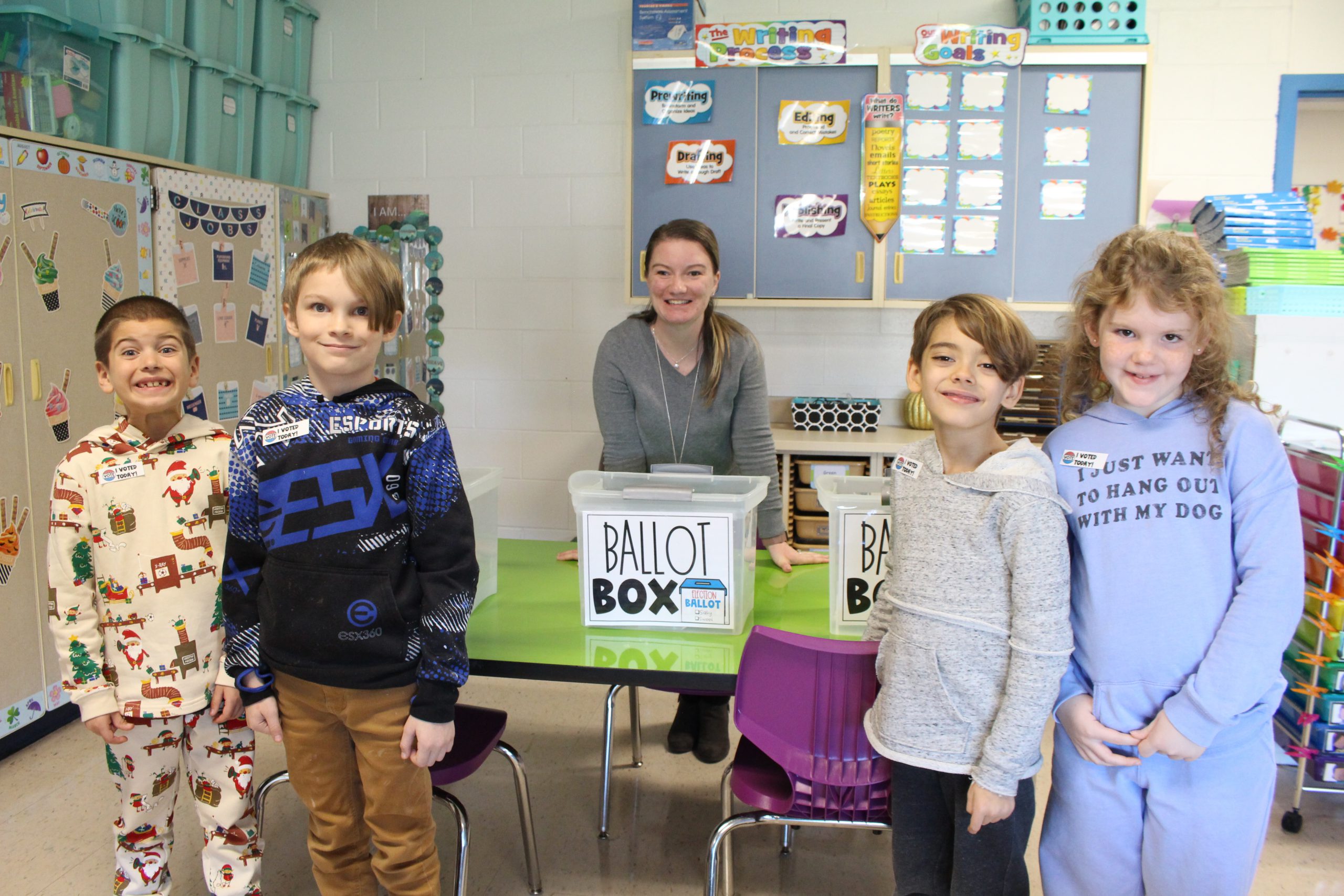 four students are standing with their teacher and smiling. In the middle there is a box with a sign on it that reads "ballot box"