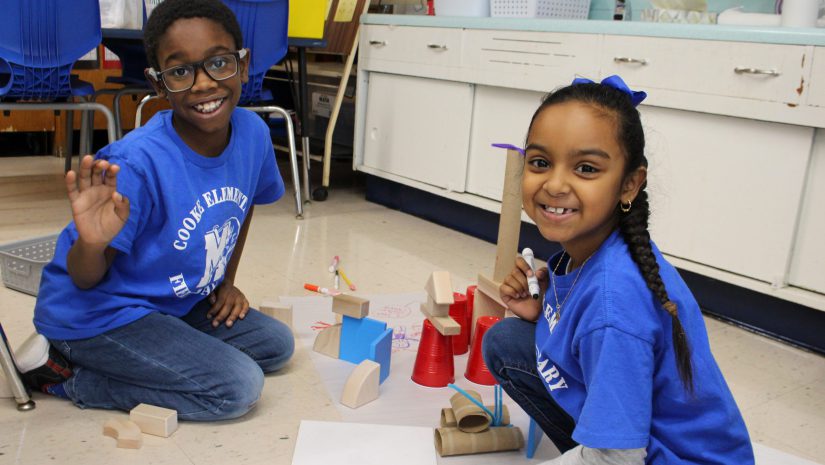 two students are drawing on a sheet of paper on the floor of a classroom. They are looking up at the camera and smiling.