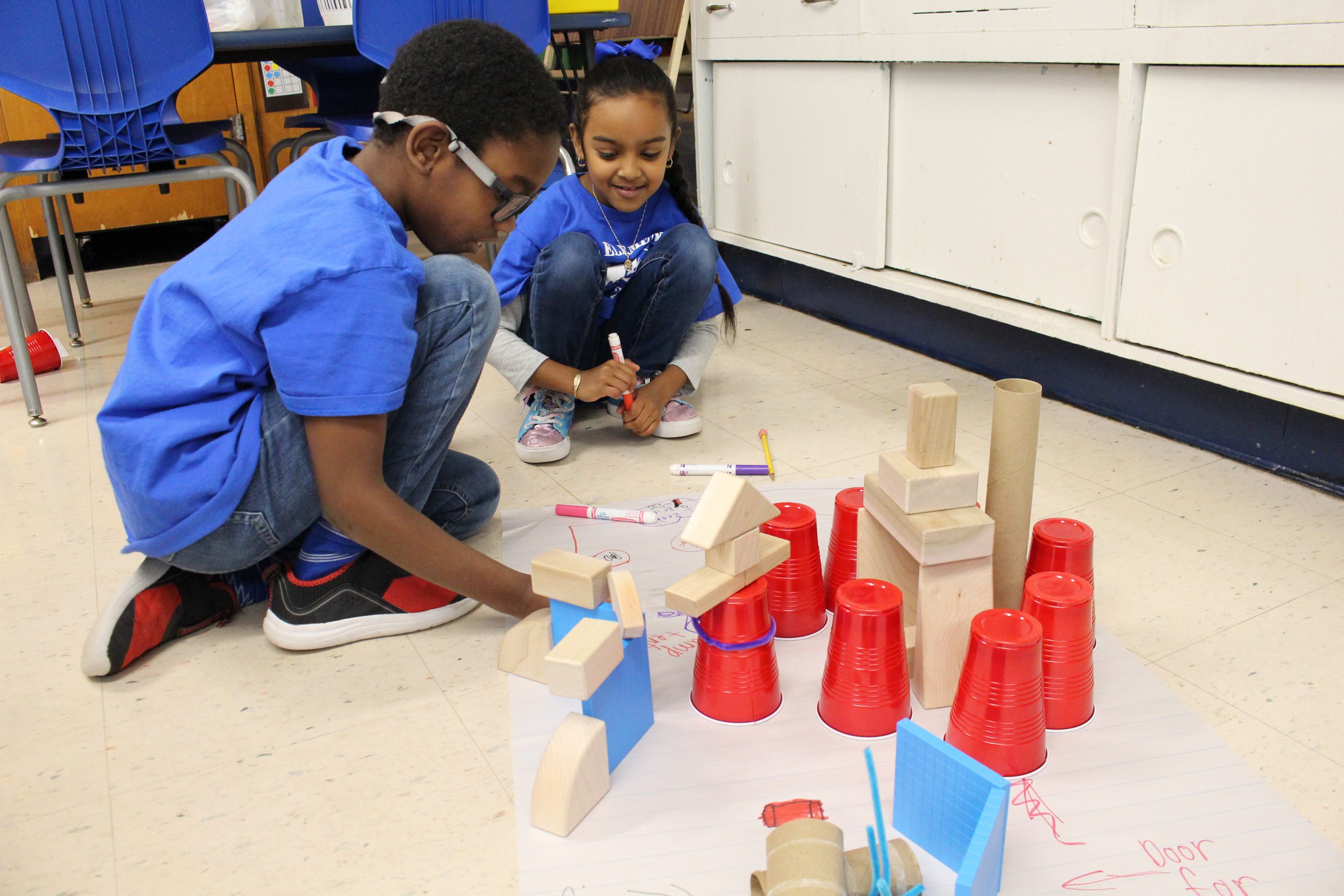 two students are crouched down on the floor and looking at a project they are working on with building blocks 