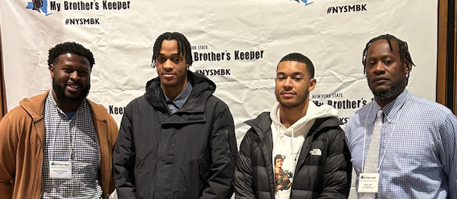 four men are posing by a backdrop that reads "MBK"