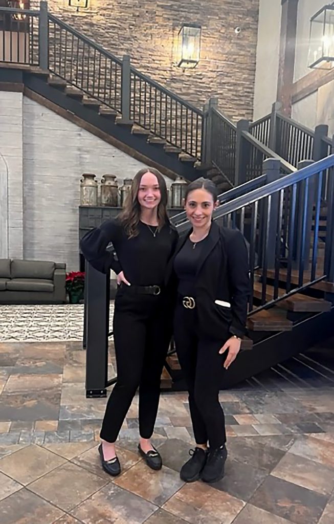 Two women standing together and smiling in front of a staircase.