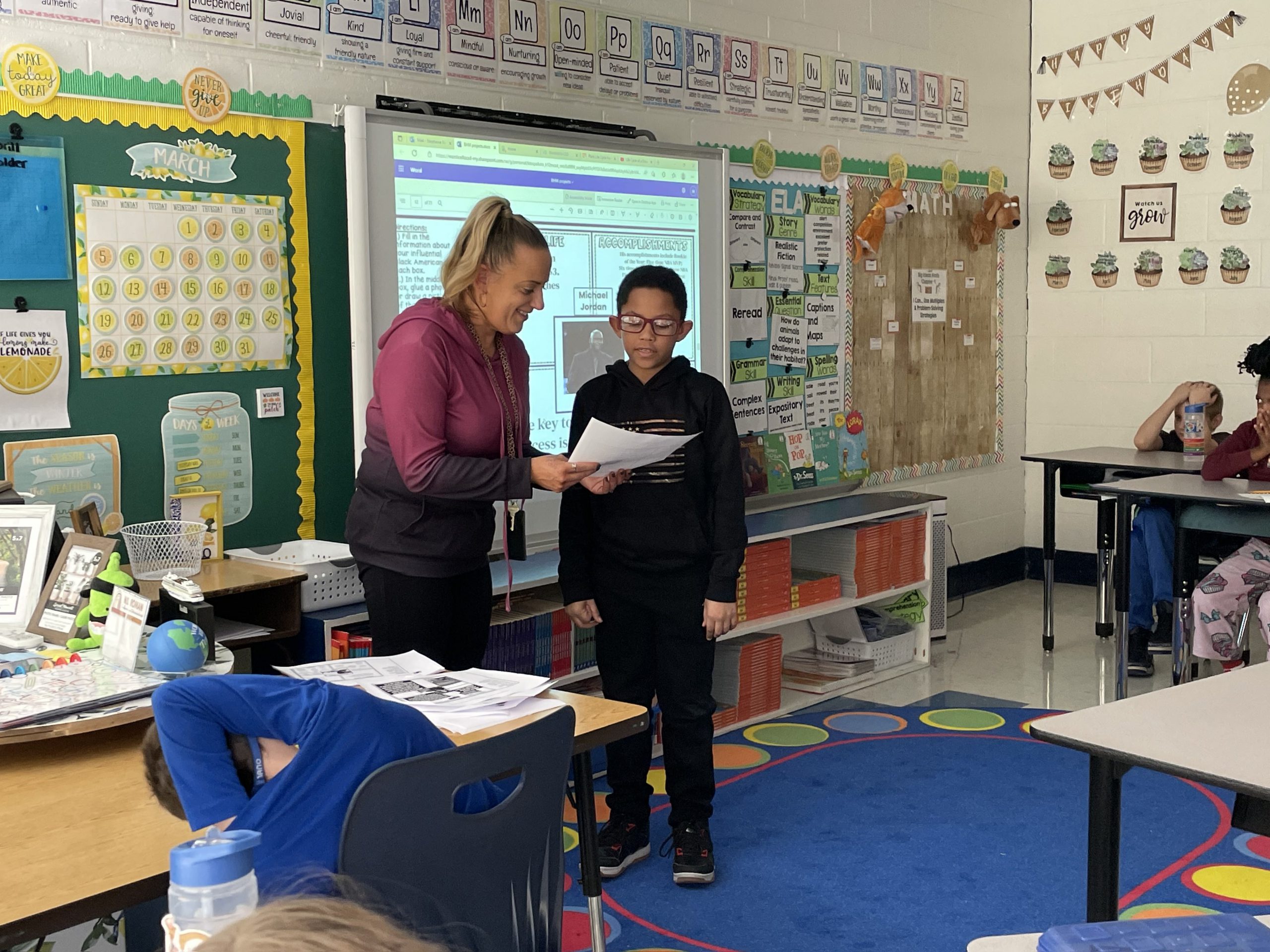 a teacher and student are standing at the front of a classroom. The student is reading off of a paper as his teacher looks on. 